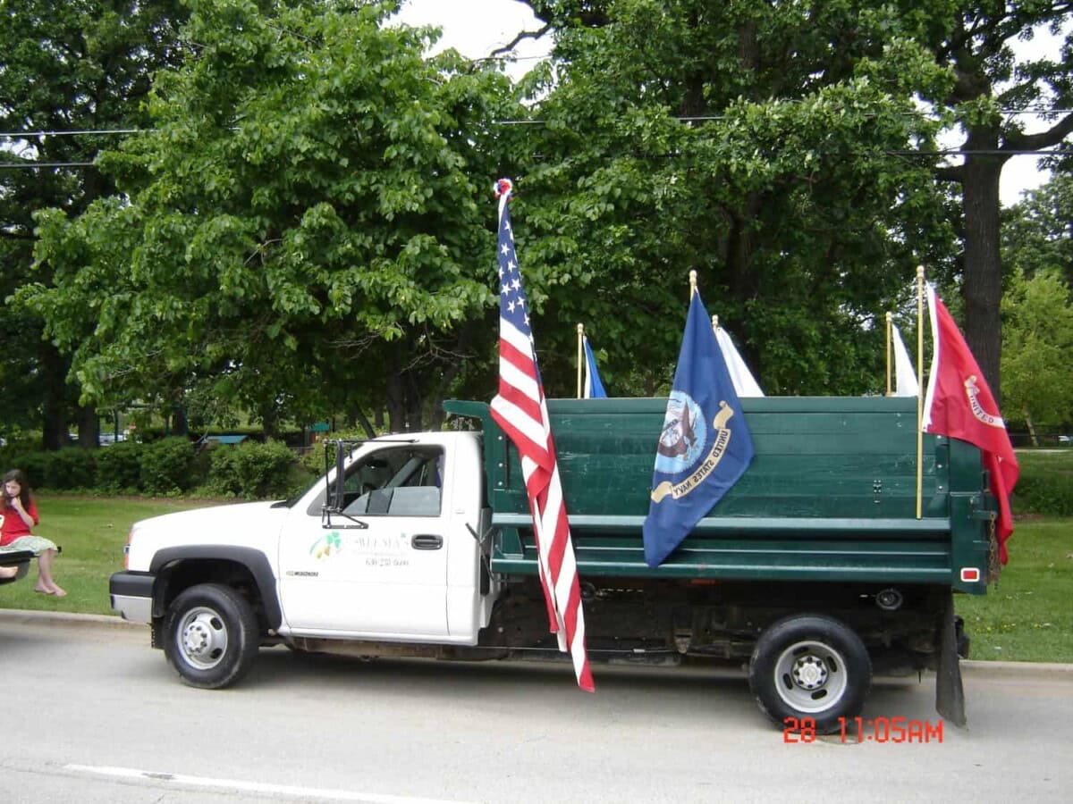 A white pickup truck with three flags, including the U.S. flag, parked on a street next to trees. A person in red sits on the grass nearby.