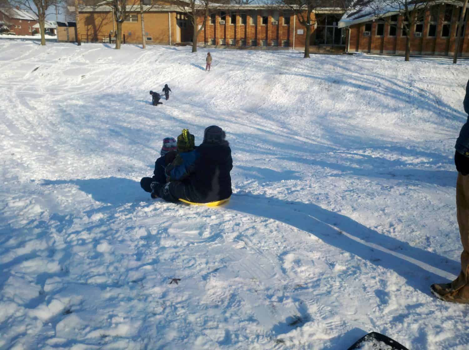 Children sled down a snowy hill on a sunny day with a brick building and scattered trees in the background.