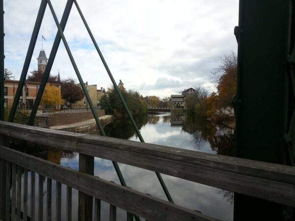 View from a wooden bridge over a calm river, with buildings, trees, and a clock tower in the background under a cloudy sky.