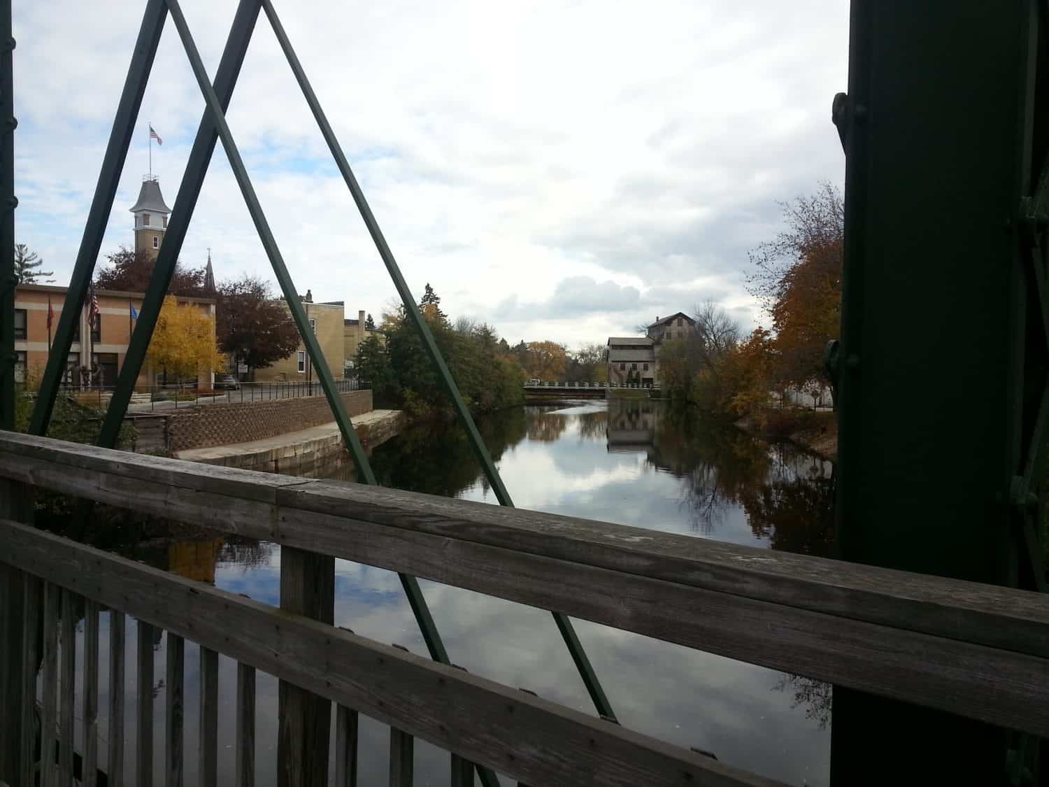 View from a wooden bridge over a calm river, with buildings, trees, and a clock tower in the background under a cloudy sky.