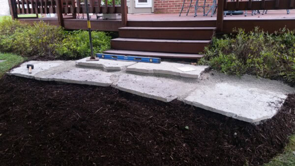 Uneven flagstone slabs are being installed on a leveled sand or gravel bed in front of a wooden porch. Tools like a level and a trowel are visible, with mulch and shrubs surrounding the work area.