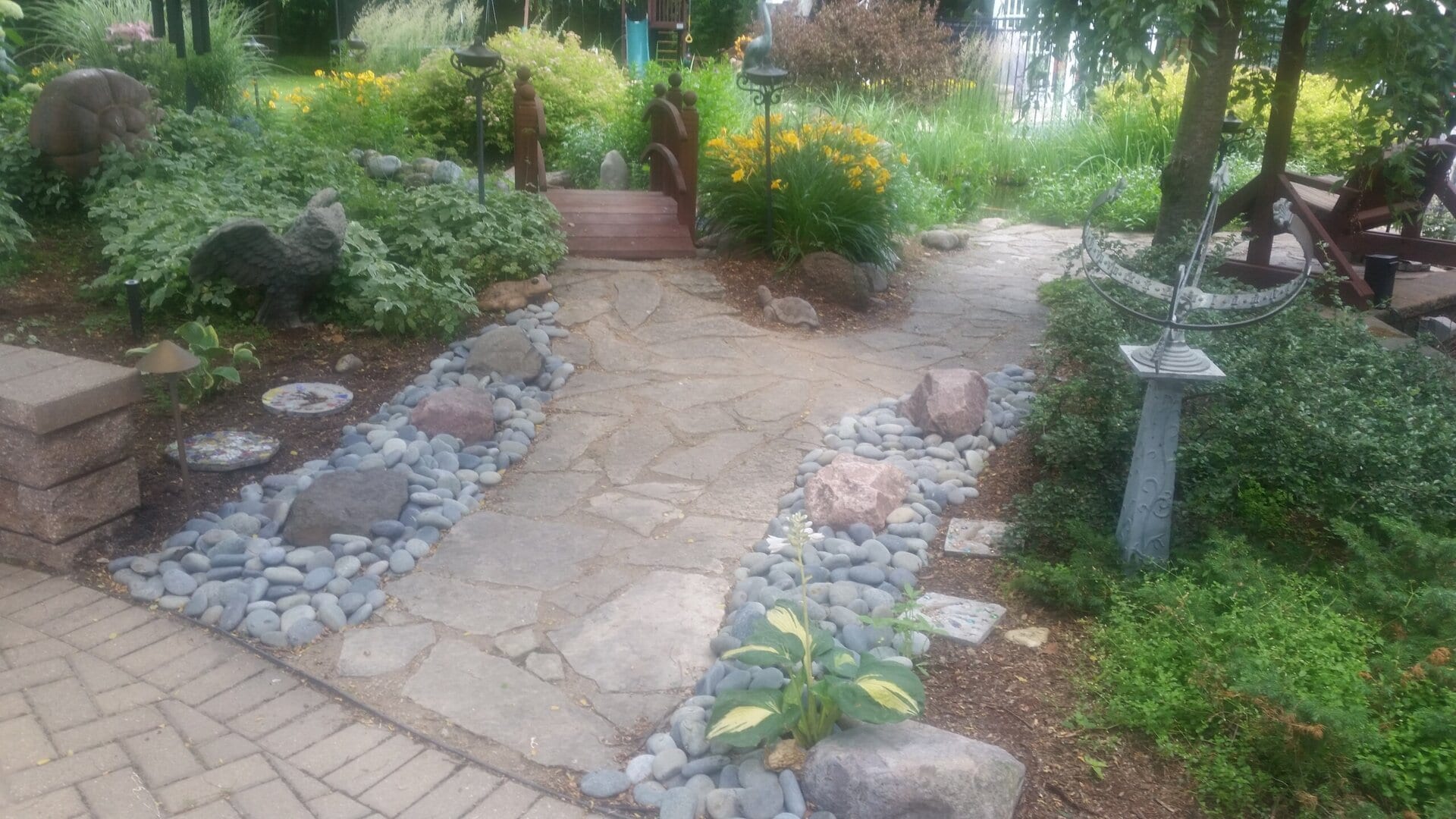 A stone path lined with gray rocks leads to a small wooden bridge in a lush garden with yellow flowers, greenery, garden statues, and decorative sundial.