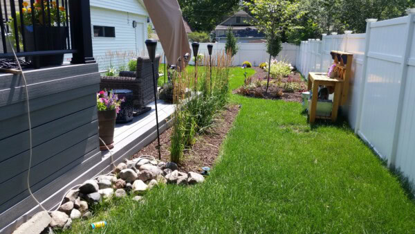 A backyard with green grass, garden beds, potted flowers, a small wooden bench, white fence, patio furniture, and rocks near a deck on a sunny day.