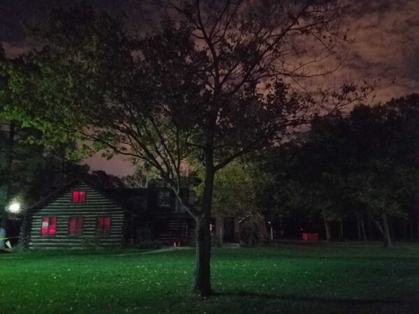 A log cabin with glowing red windows sits among trees at night, illuminated by greenish outdoor lighting. The sky is cloudy, casting an eerie atmosphere over the scene.