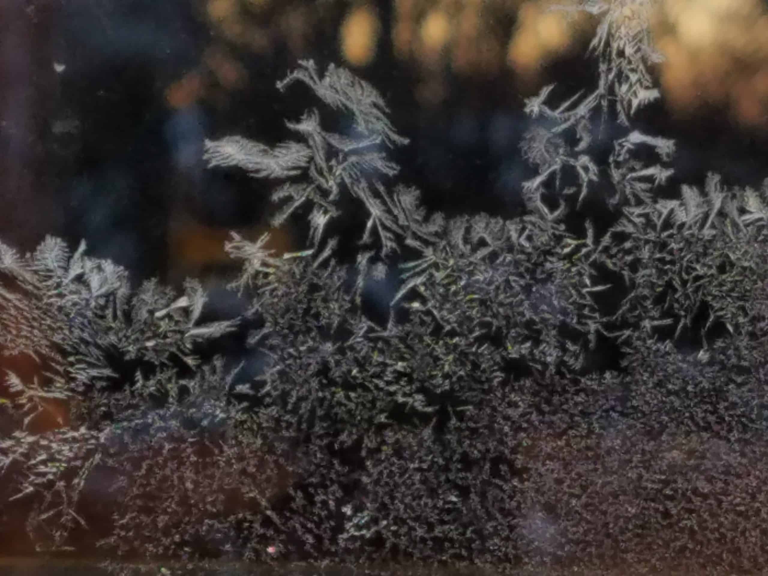 Close-up of frost crystals forming intricate patterns on a window, with a blurred background showing hints of light and dark colors.