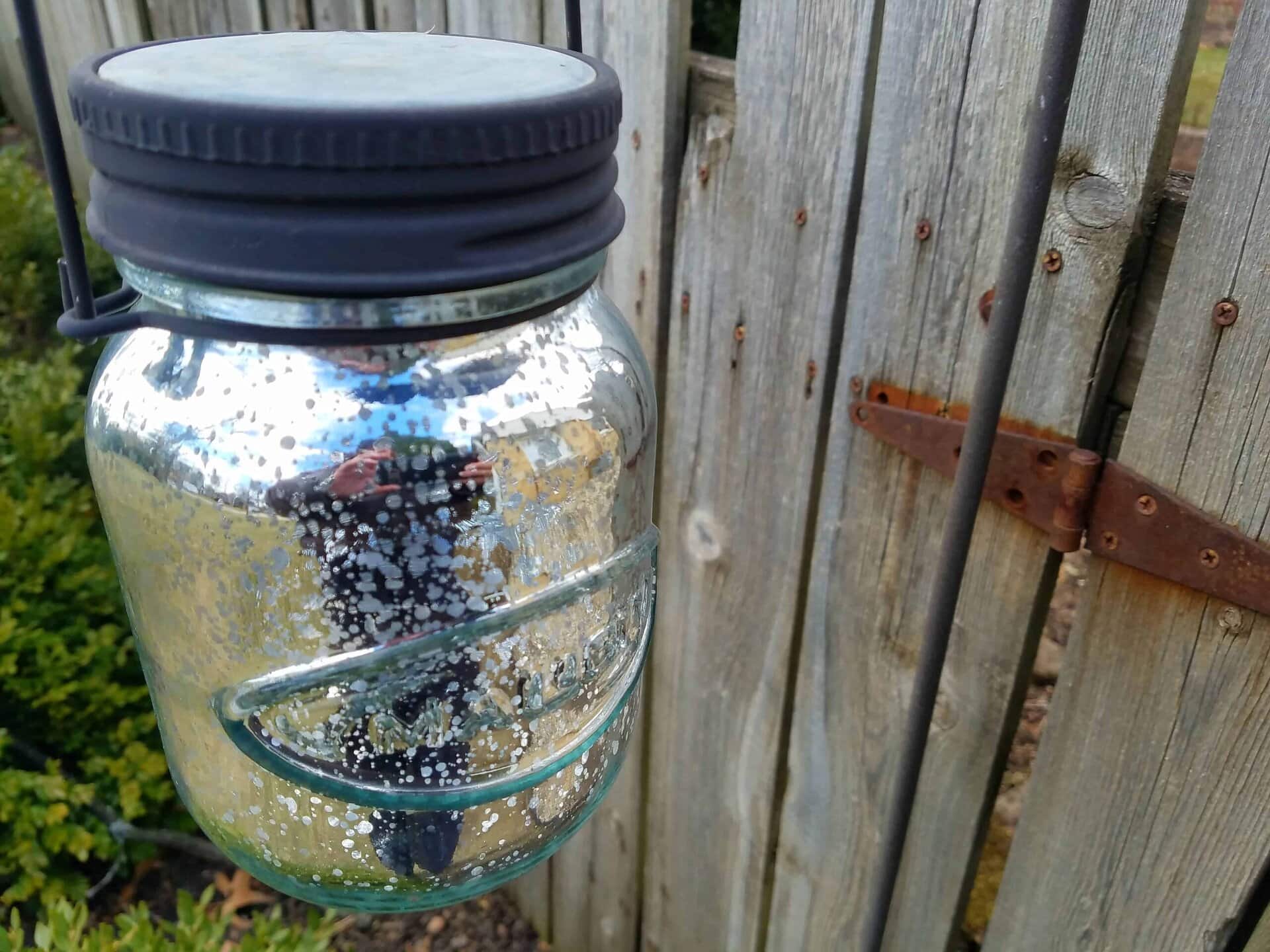 A speckled glass jar with a black lid hangs from a metal handle in front of a weathered wooden fence with a rusty hinge; greenery is visible in the background.