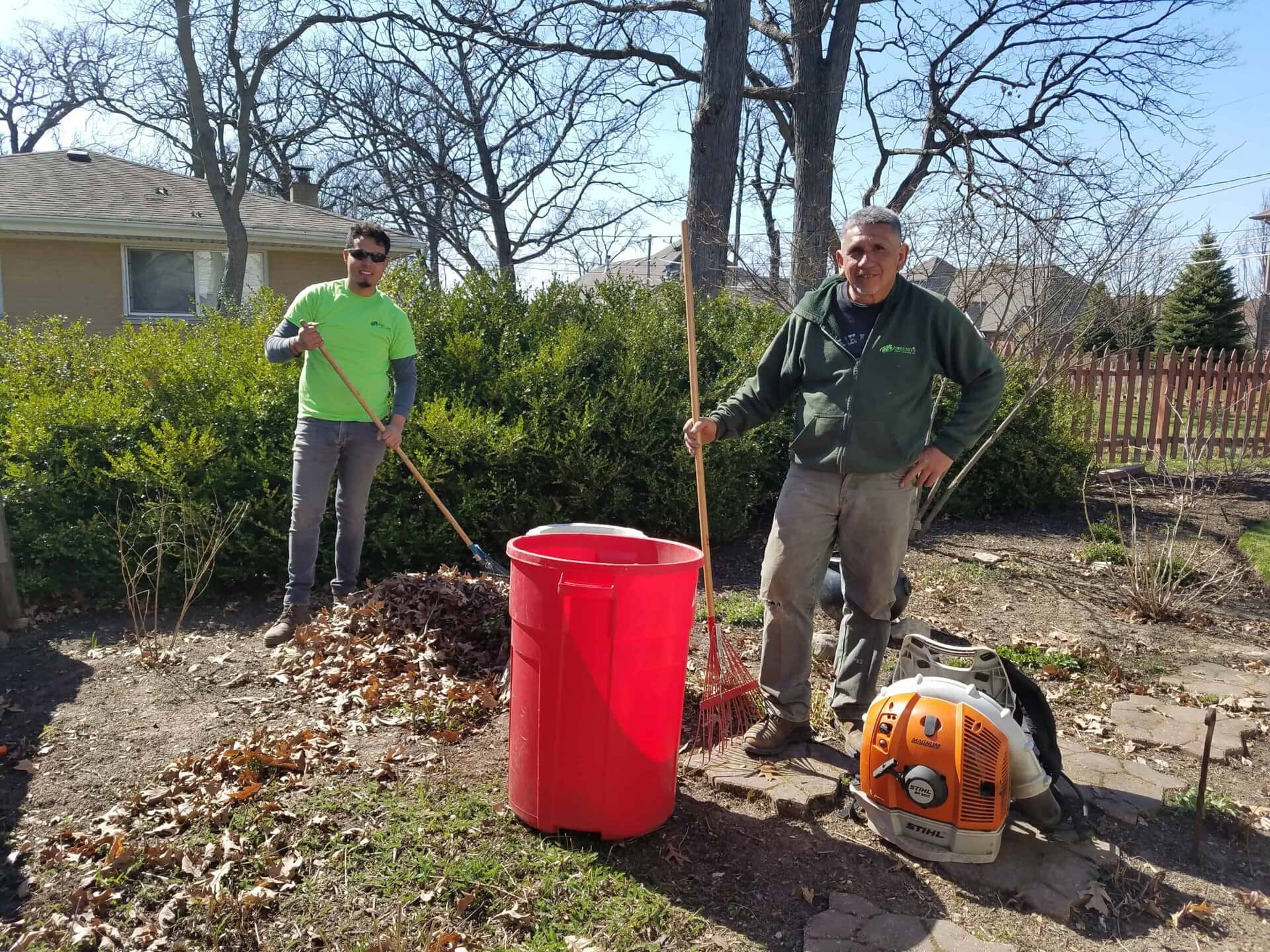 Two men raking dry leaves in a yard on a sunny day. One man holds a rake and stands by a red bin, while the other man rakes leaves into a pile. Leaf blower on ground. Bushes and trees in background.