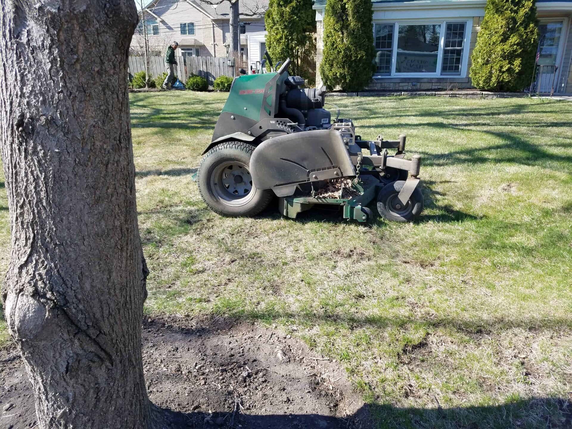 A large green lawn mower sits on patchy grass near a tree in a residential yard, with houses and shrubs in the background.