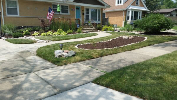 A neatly landscaped front yard with a mulched garden bed edged with rocks, a green lawn, a U-shaped sidewalk, American flag, small shrubs, and two houses in the background.