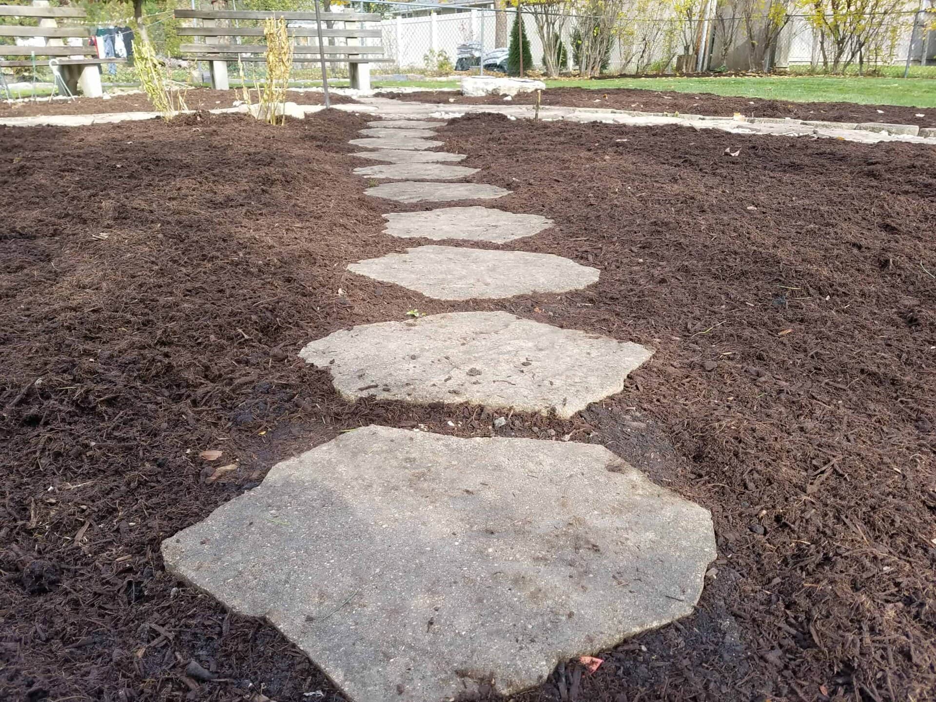 A row of flat, hexagonal stepping stones forms a path through dark mulch in a garden, with benches and plants in the background.
