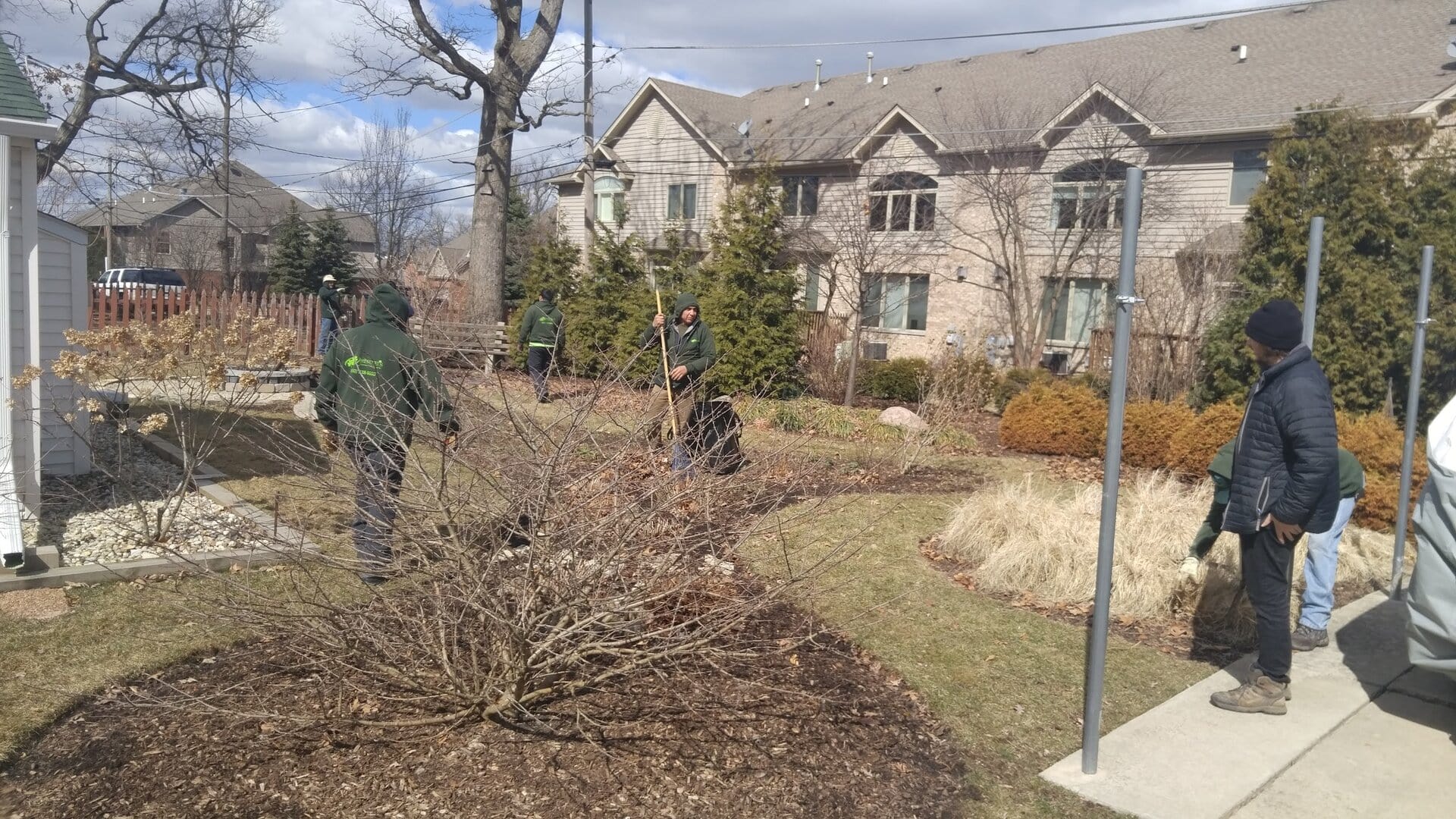 Four people in outdoor clothing stand and work in a residential garden on a cloudy day. Leafless shrubs and ornamental grasses fill the yard, and houses are visible in the background.