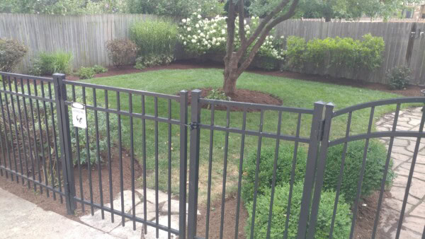 Black metal gate in the foreground opens to a well-maintained backyard with green grass, shrubs, a tree surrounded by mulch, and a wooden fence enclosing the area. A sign on the gate shows a dog silhouette.