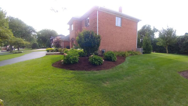 A red-brick house with neatly trimmed green lawn, shrubs, and mulched flower beds in a suburban neighborhood on a sunny day.
