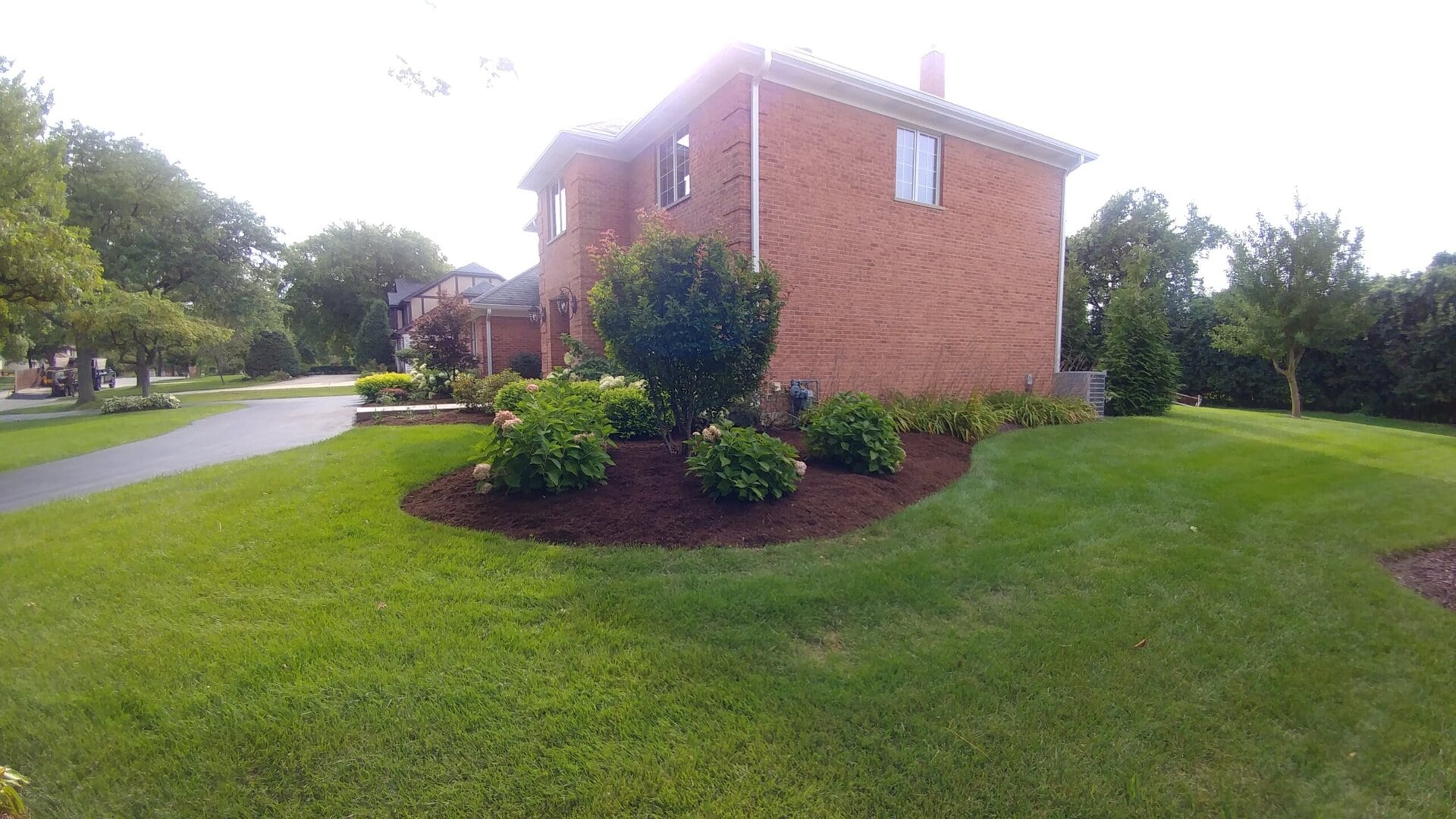 A red-brick house with neatly trimmed green lawn, shrubs, and mulched flower beds in a suburban neighborhood on a sunny day.