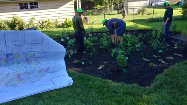 Three people plant young plants in a garden bed while referencing a hand-drawn garden plan on graph paper in the foreground. The area is fenced, with houses and grass visible in the background.