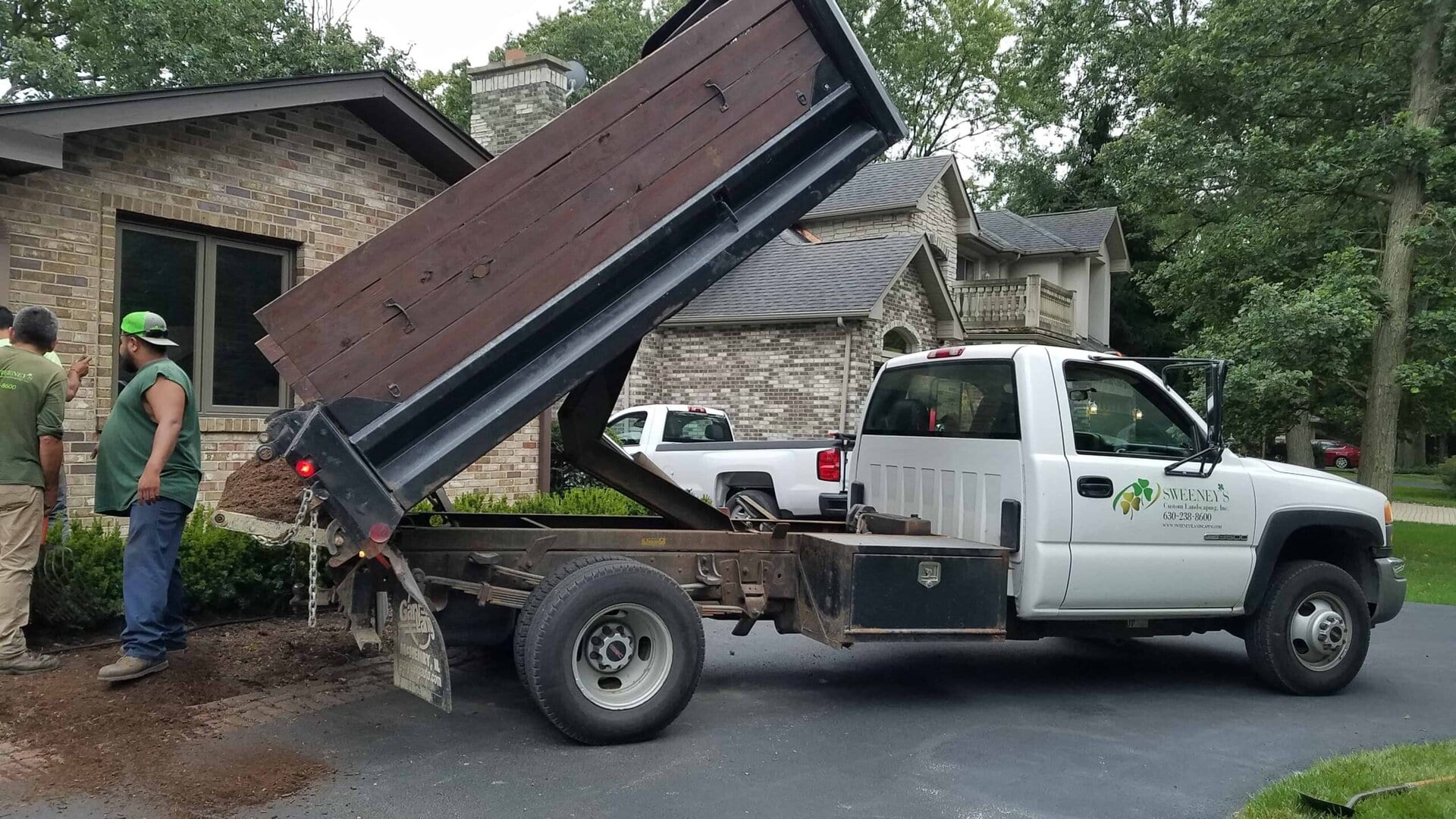 A dump truck unloads soil onto a driveway next to a brick house while two workers stand nearby, preparing for landscaping work. Trees and another vehicle are visible in the background.