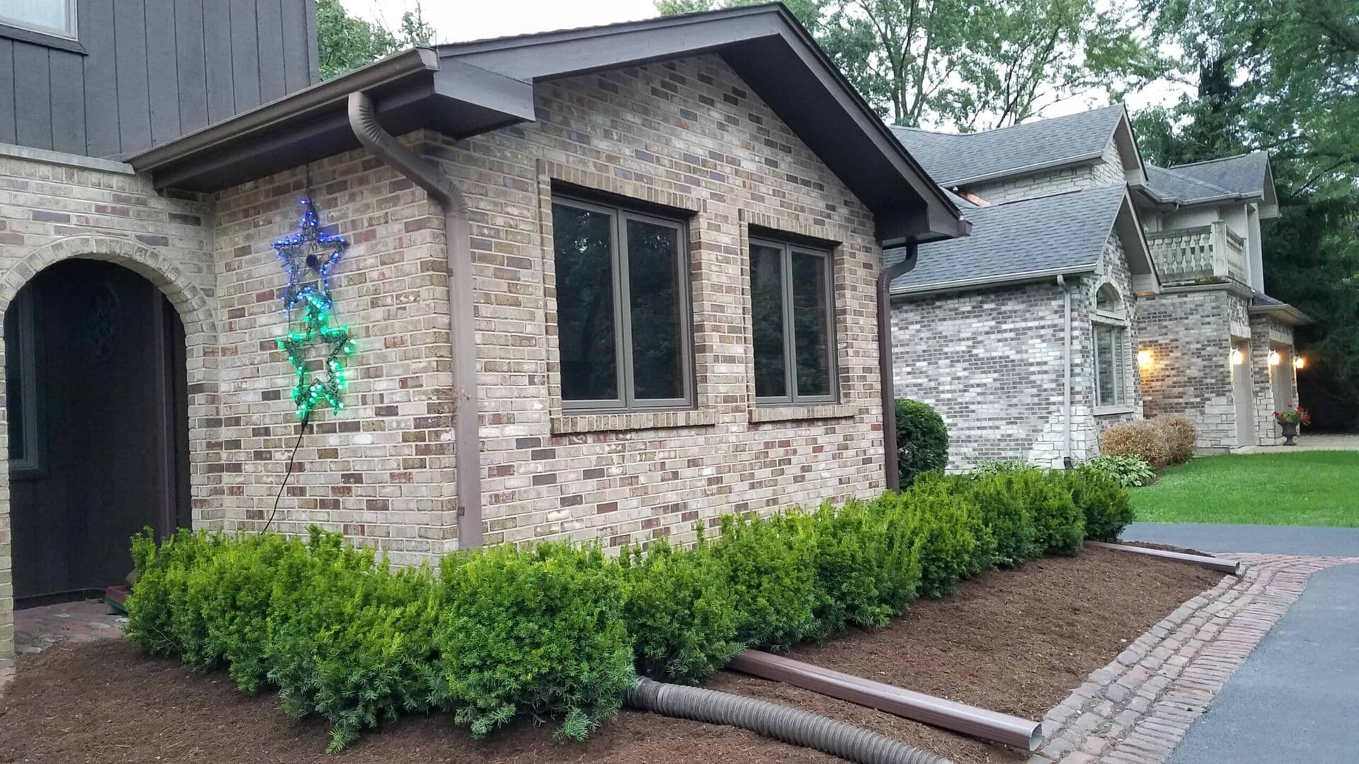 A brick house with green shrubs, star-shaped decorations on the wall, and a black drainage pipe extending into a mulched garden bed near a driveway.