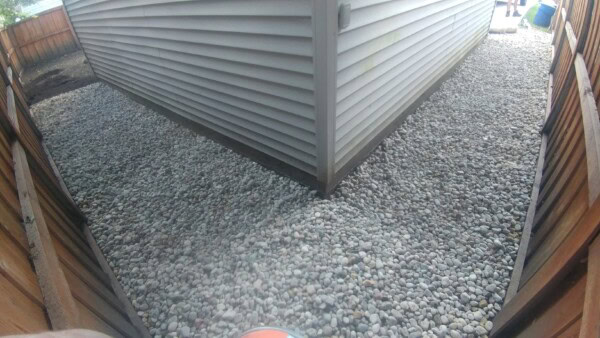 A corner of a house with gray siding is surrounded by a yard covered in small gray rocks, bordered by a wooden fence.