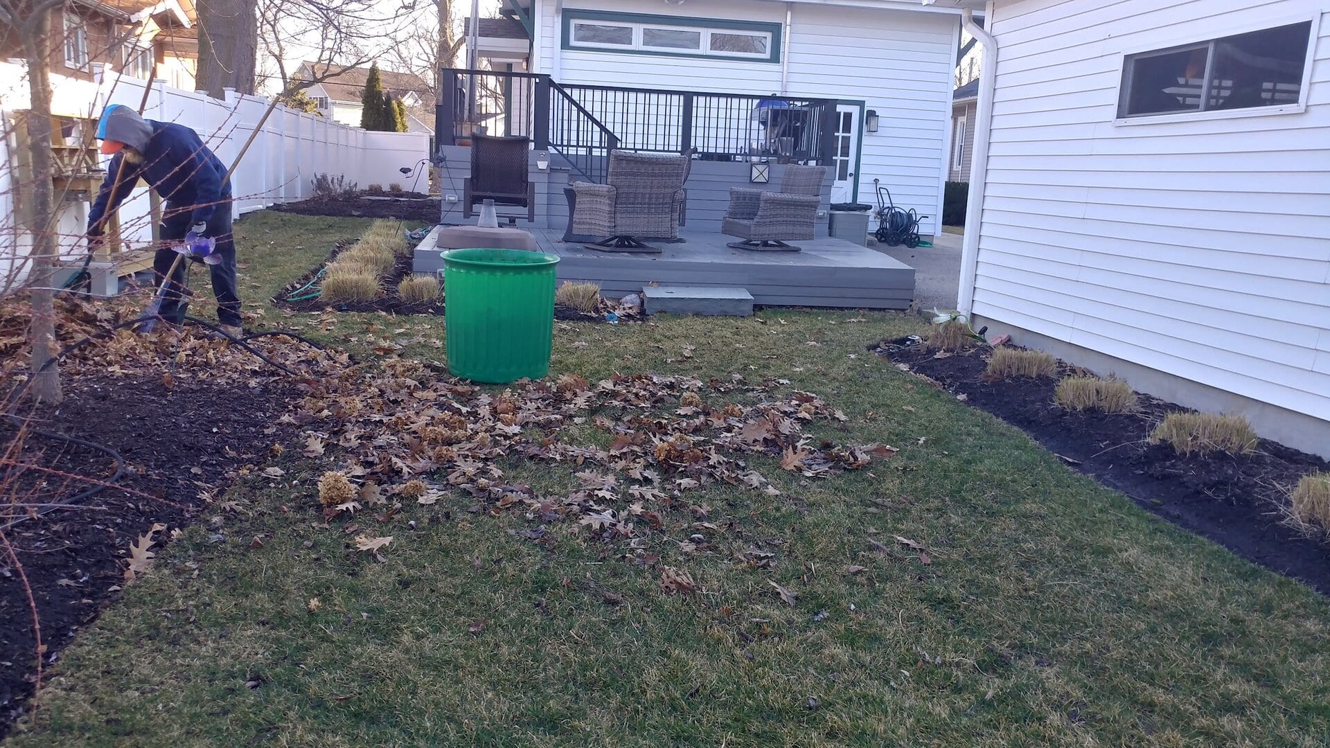 A person rakes fallen leaves into a green bin in a backyard with a deck, patio chairs, and garden beds, beside a white house.