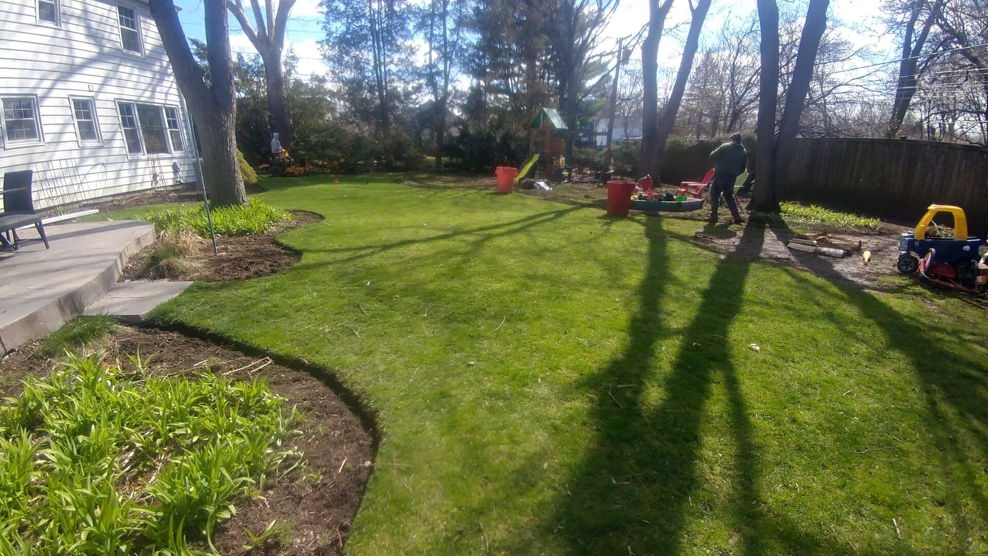 A backyard with green grass, garden beds, trees casting long shadows, and a person working near red containers; a childs play area is visible in the distance.