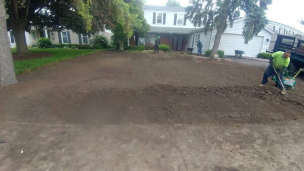 A person operates a green rototiller on freshly turned soil in front of a suburban house, preparing the yard for landscaping. Trees frame the scene, and the driveway is visible on the left.