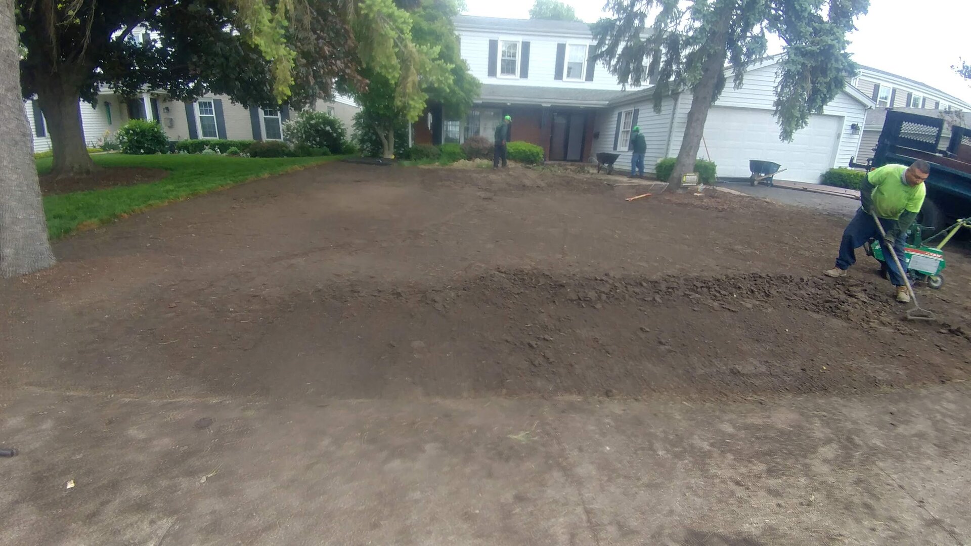 A person operates a green rototiller on freshly turned soil in front of a suburban house, preparing the yard for landscaping. Trees frame the scene, and the driveway is visible on the left.