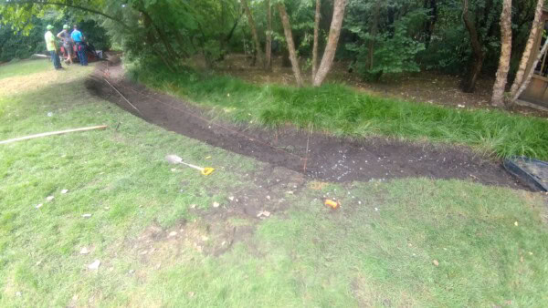 A curved trench is dug in grassy ground with string lines marking its borders. Trees and bushes line the background, and a few people stand in the distance. Tools are scattered near the trench.