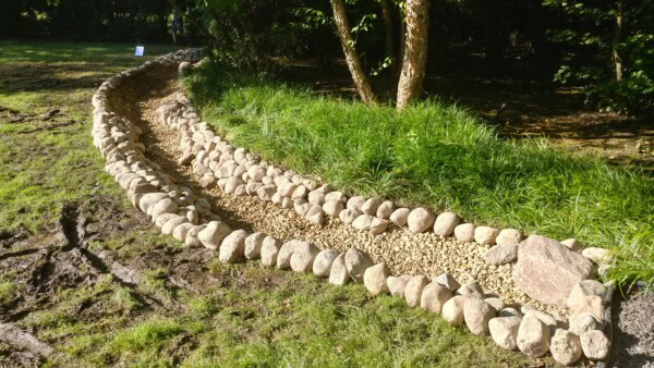 A dry creek bed lined with medium and large round stones winds through grassy and shaded areas, bordered by taller grass and trees.