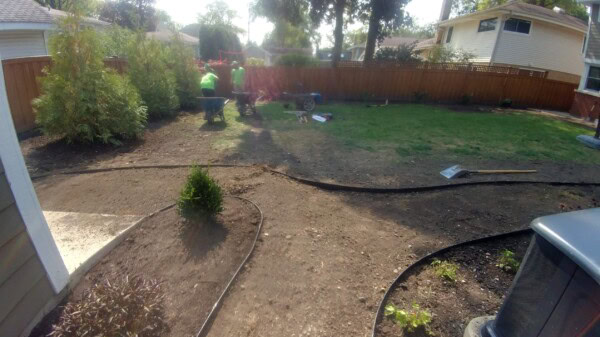 Three people work in a backyard, using wheelbarrows and tools. The yard is partially dug up with soil and some grass remaining. There are bushes, a wooden fence, and two houses visible.