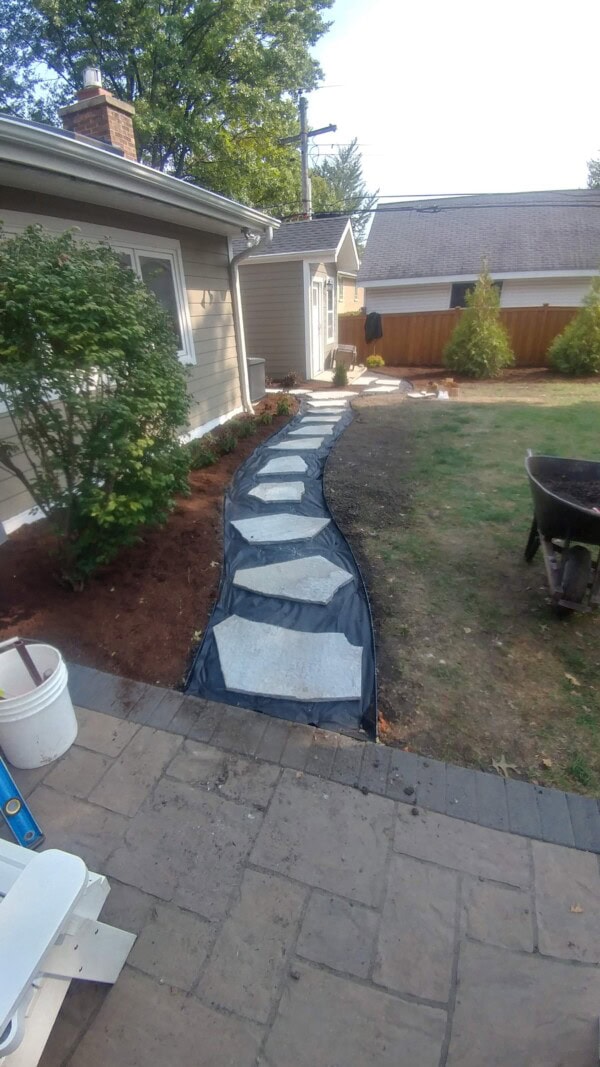 A stone pathway is being constructed alongside a house, with stones placed on top of black landscaping fabric and brown garden mulch lining the edge. Gardening tools and a wheelbarrow are nearby.