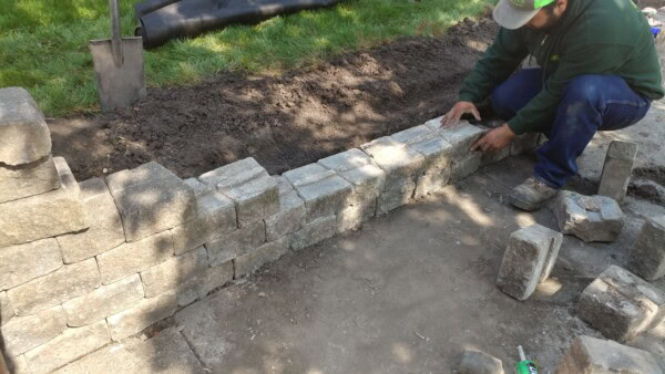 A person in work clothes is building a low stone wall, carefully placing rectangular stones. Fresh soil and green grass are visible in the background, and loose stones and a shovel are nearby.