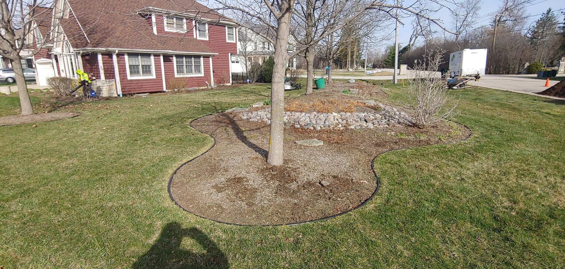 A tree stands in a mulched garden bed with stone edging on a grassy lawn. A red house and another landscaped area are in the background under a clear sky. Shadows and landscaping tools are visible.