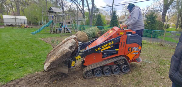 A person operates an orange mini skid steer to move a large tree root ball in a backyard near a green fence and a playset on a cloudy day.