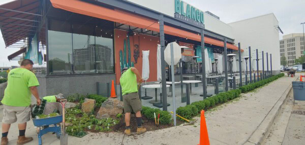 Three workers in neon shirts landscape the area outside a restaurant with outdoor seating, placing plants and rocks near the sidewalk. Orange cones and tools are scattered around the site.