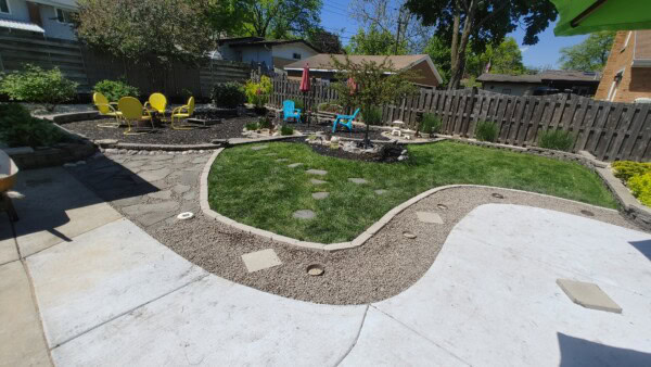 Backyard with green grass, curved gravel paths, yellow and blue chairs around two separate fire pits, and a fence with trees and shrubs along the border on a sunny day.
