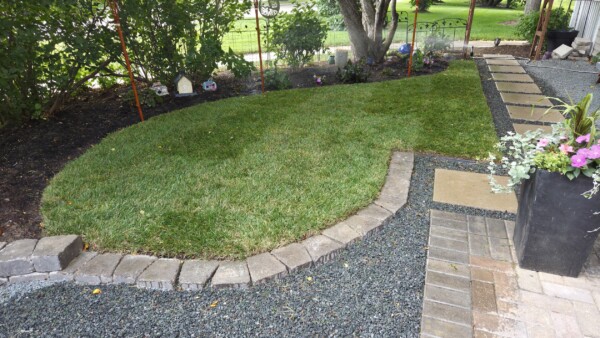 A small, neatly edged lawn is bordered by stone bricks, gravel paths, and a flower bed with blooming plants. A potted plant with pink flowers sits near stone pavers leading to a house.