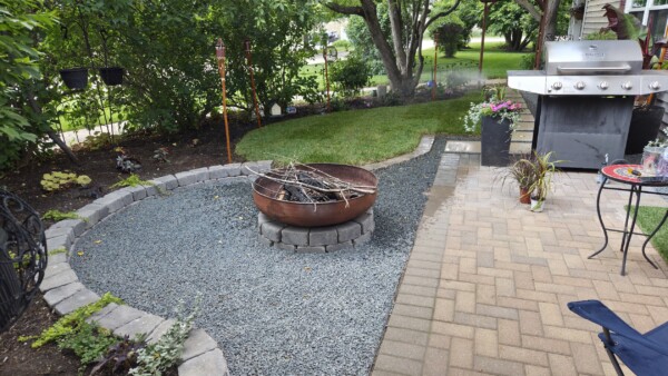 A backyard with a round stone fire pit on gravel, surrounded by garden beds, a brick patio, a stainless steel grill, and potted plants, under leafy trees.