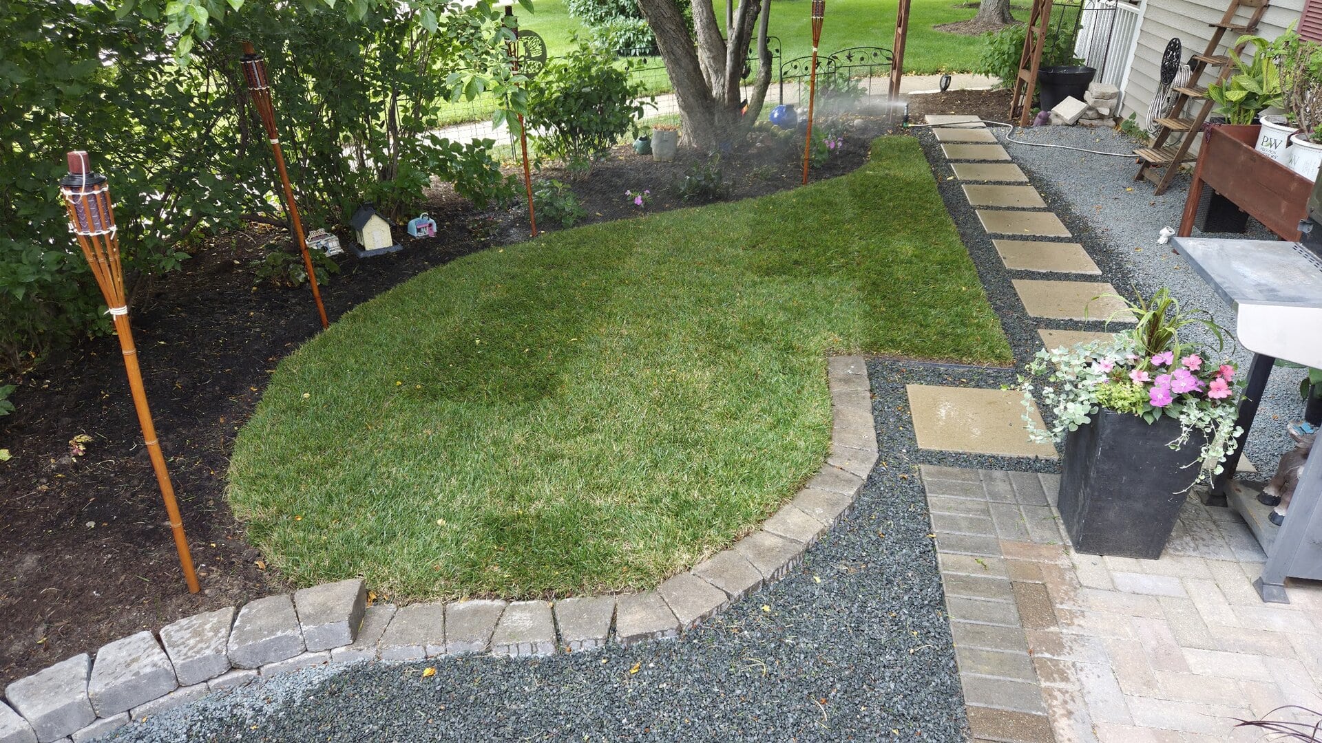 A small backyard garden with a curved green lawn, tiki torches, paver stone paths, a flower planter, mulch beds, a barbecue grill, and outdoor chairs under a leafy tree.