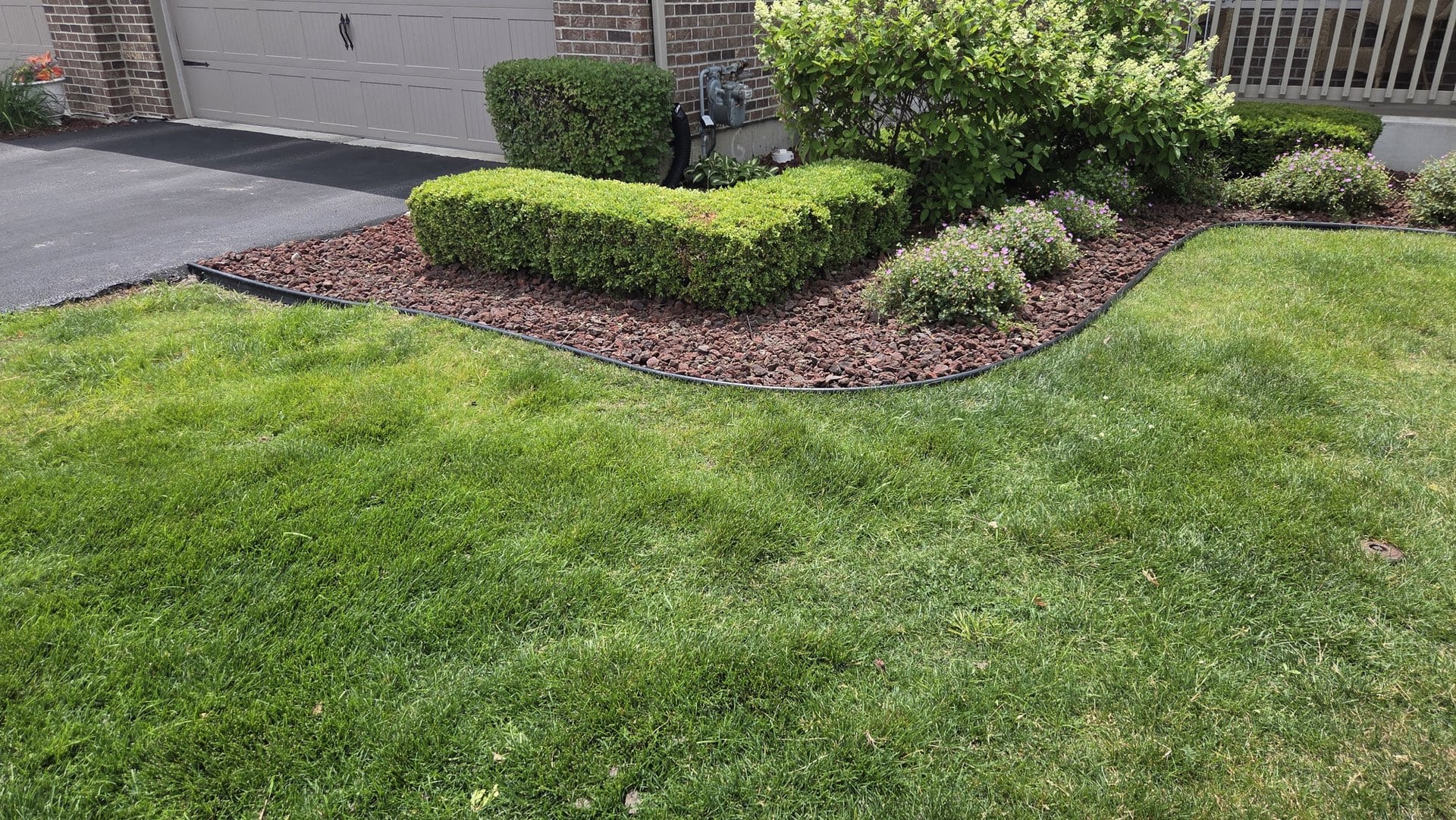 A neatly landscaped yard with green grass, trimmed bushes, small flowering plants, and red mulch next to a driveway and garage.