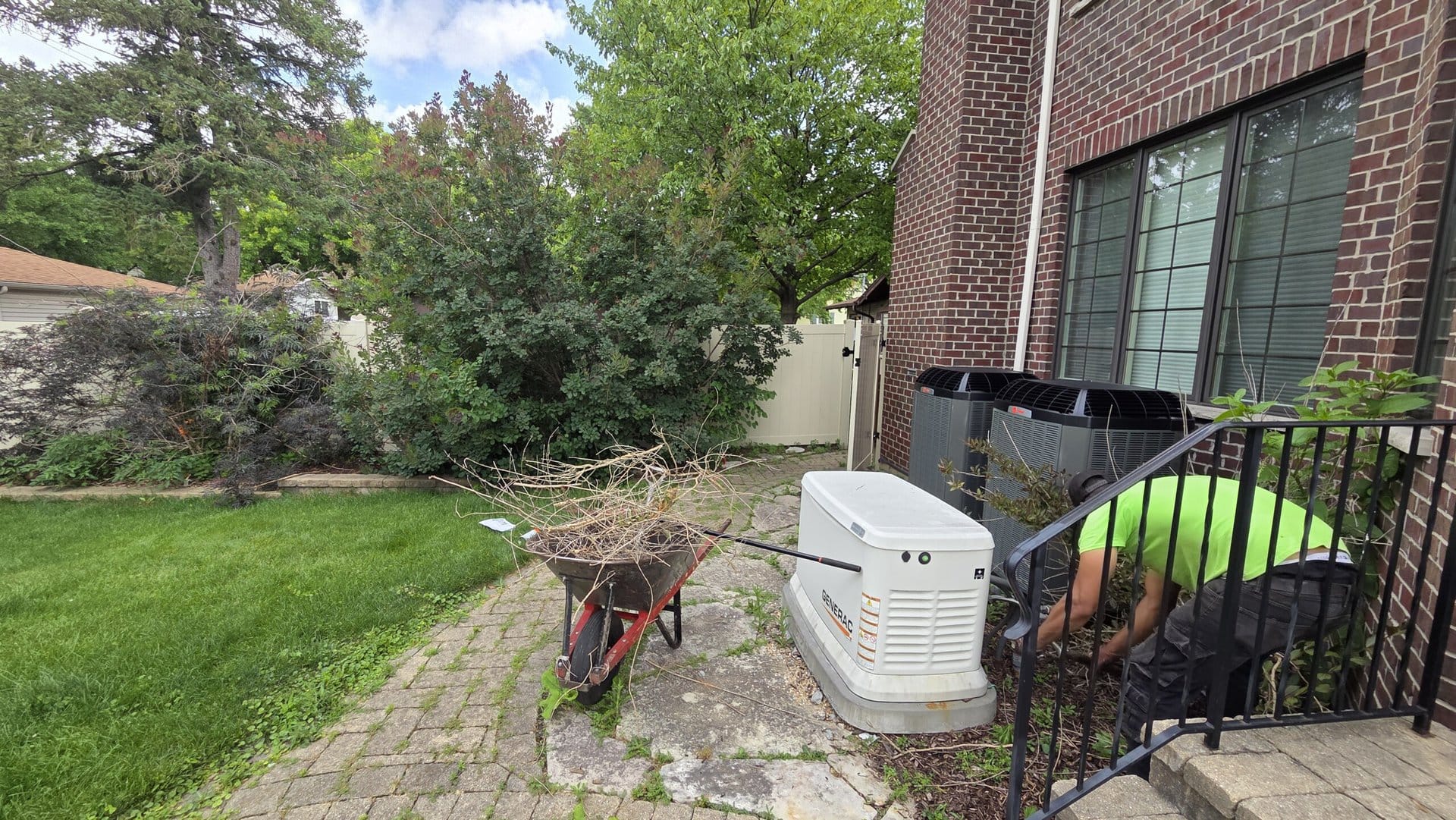 A person in a green shirt works near a generator and wheelbarrow filled with branches beside a brick house with large windows. Lush greenery and a lawn are visible in the background.