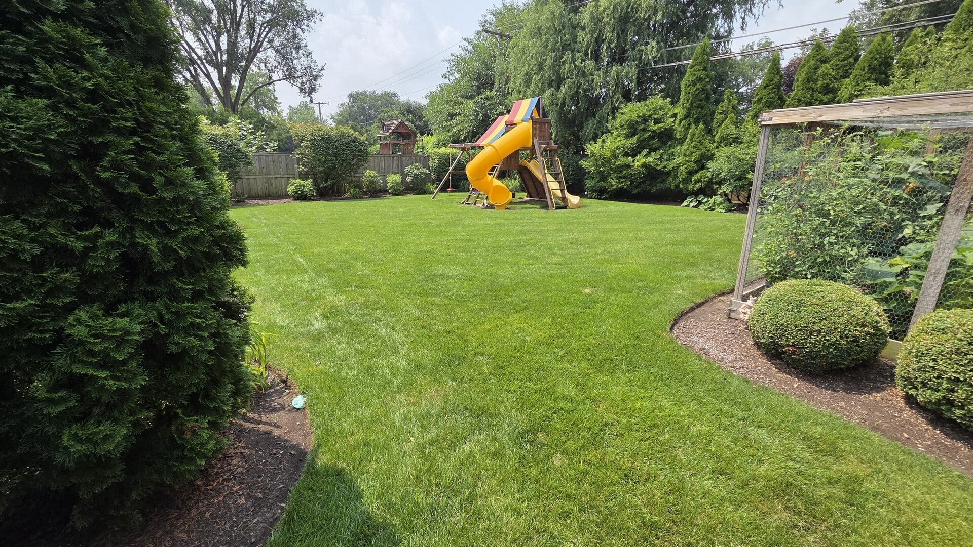 A backyard with a green lawn, a playset featuring a yellow slide, trees, bushes, and a fenced garden on the right side.