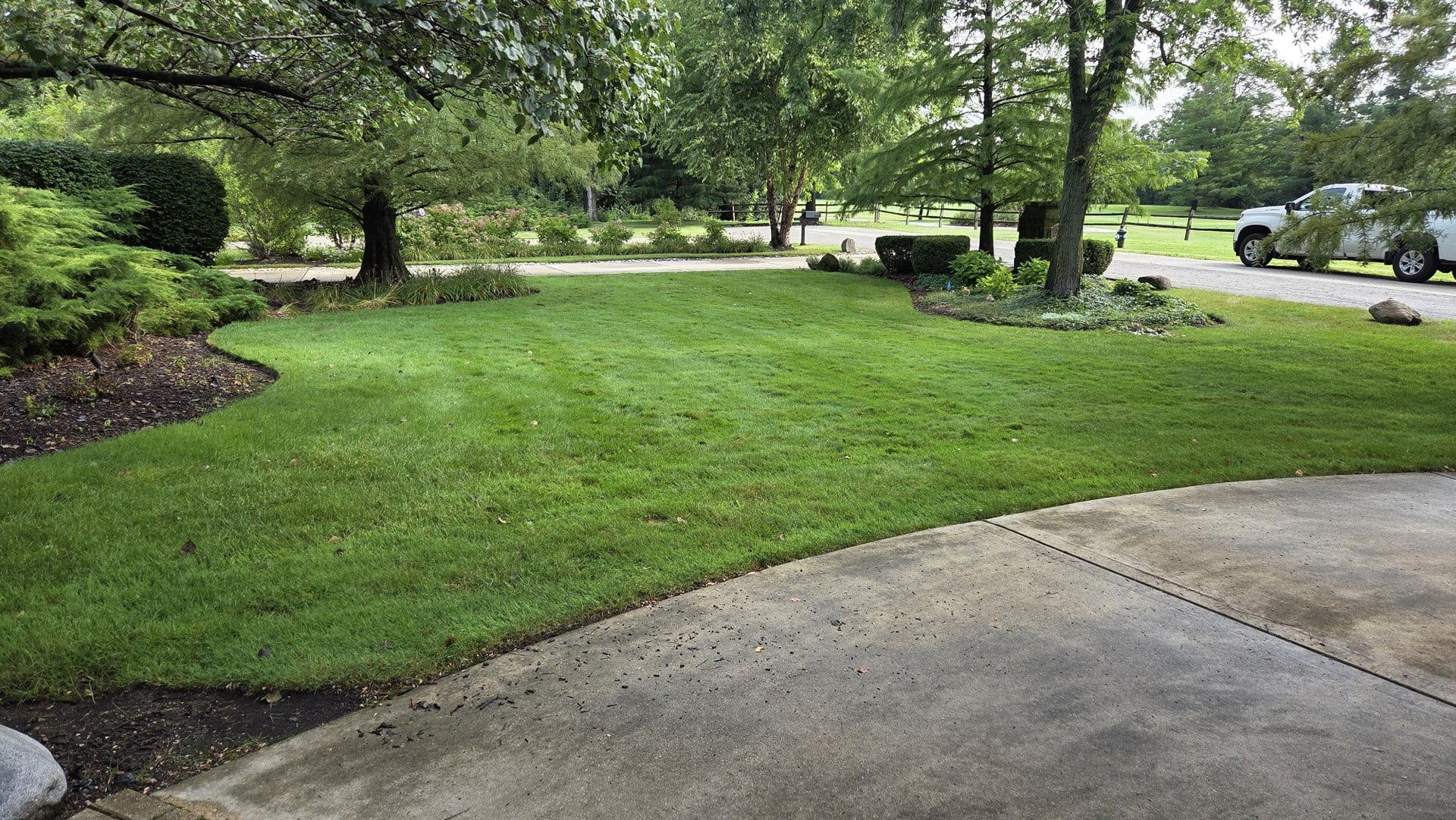 A well-kept lawn with neatly trimmed grass, bordered by trees, shrubs, and a curved concrete driveway. A white vehicle is parked in the distance near a quiet, tree-lined road.