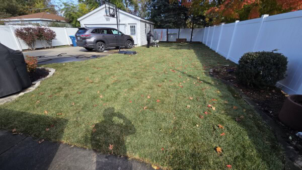A fenced backyard with a patchy lawn, scattered autumn leaves, a white garage, parked car, and driveway. A persons shadow is visible in the foreground, with trees and shrubs along the sides.
