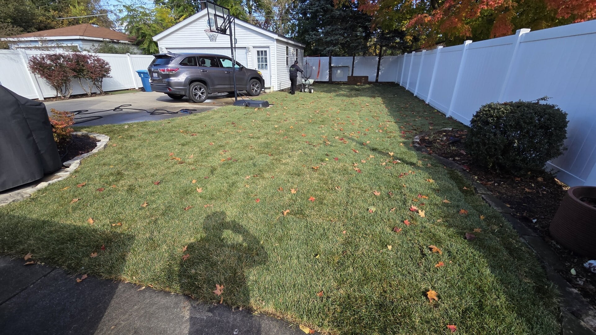 A fenced backyard with a patchy lawn, scattered autumn leaves, a white garage, parked car, and driveway. A persons shadow is visible in the foreground, with trees and shrubs along the sides.