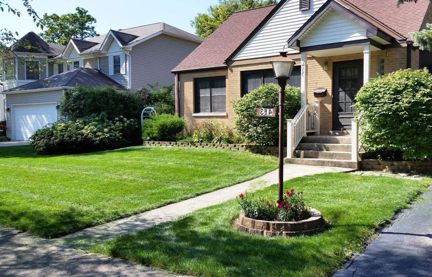 A well-kept suburban house with a manicured lawn, trimmed bushes, and a black lamp post by the sidewalk. Steps lead to the front door, and a white garage is visible next door.