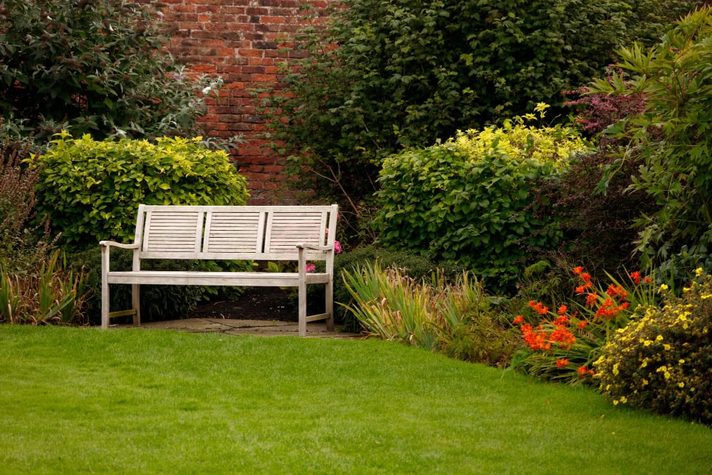 A wooden bench sits on a stone path in a garden, surrounded by green shrubs and colorful flowers, with a red brick wall in the background and a neatly trimmed grass lawn in the foreground.