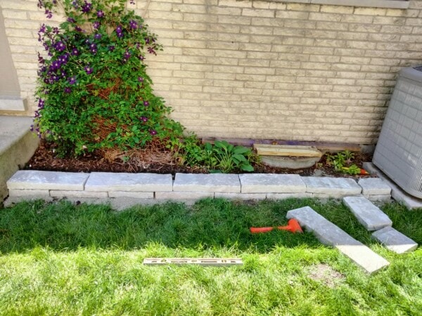 A partially built stone garden border runs along a flower bed by a brick wall, with tools and extra stones on the grass nearby. A leafy plant with purple flowers grows on the left.