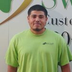 A man in a lime green shirt stands smiling in front of a sign with green and gold text and a logo. The shirt has a small company logo on the left chest.