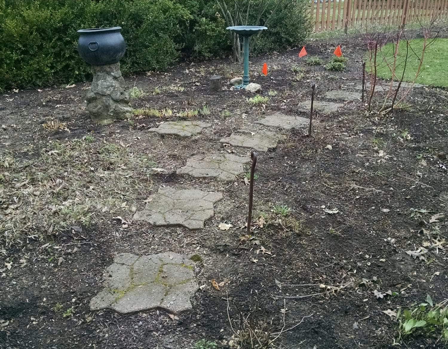 Stepping stones form a path through a garden bed with sparse plants. A black planter on a stone base and a birdbath are visible. Orange flags mark certain spots in the soil. A wooden fence stands in the background.