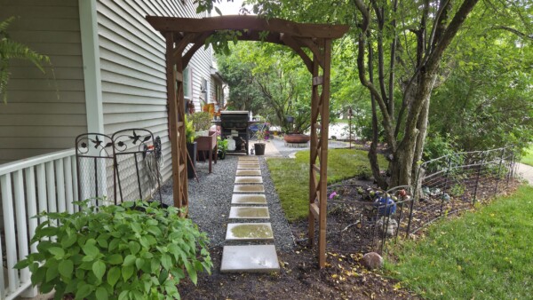 A wooden garden archway stands over a stepping stone path leading to a grill and patio area beside a house, surrounded by lush green plants and trees.